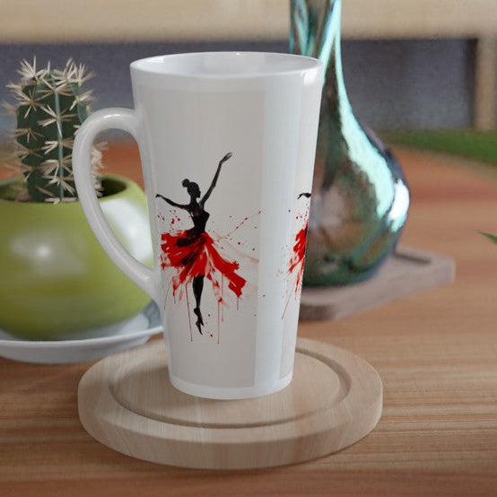 A white ceramic latte mug with a black and red ballet dancer silhouette design, resting on a round wooden coaster on a wooden table next to a small potted cactus.