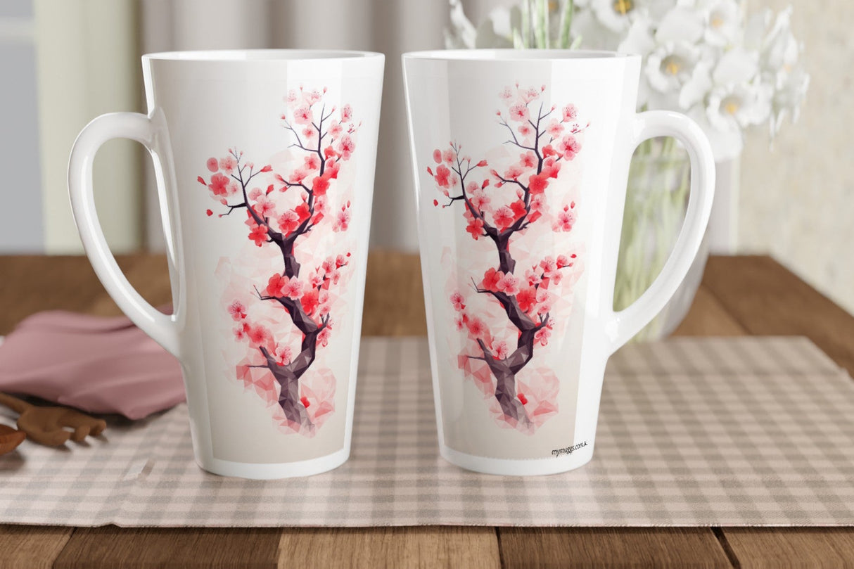 Two tall white ceramic mugs with pink and red cherry blossom branch illustrations, displayed on a checkered placemat on a wooden table with a blurred floral background.