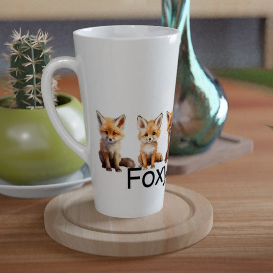 A white ceramic latte mug with a printed graphic of two foxes and the word 'FOX' sits on a round wooden coaster on a wooden table, styled with a small potted cactus and a green plant in a home setting.