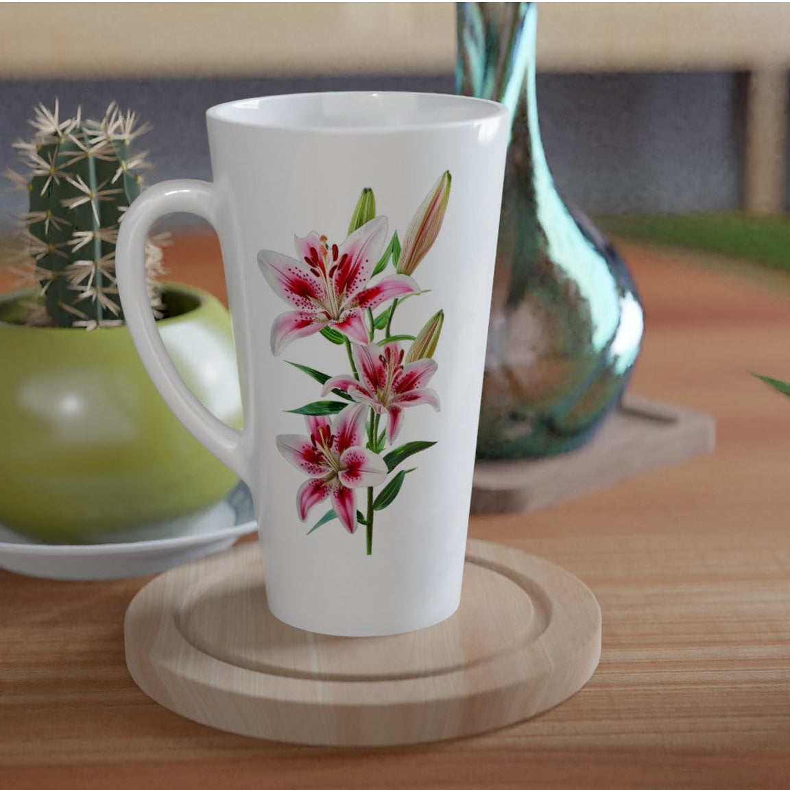 A white ceramic latte mug with a pink and green lily floral print, resting on a round wooden coaster on a wooden table. In the background, a small green cactus in a green pot and a metallic teal glass vase are visible.