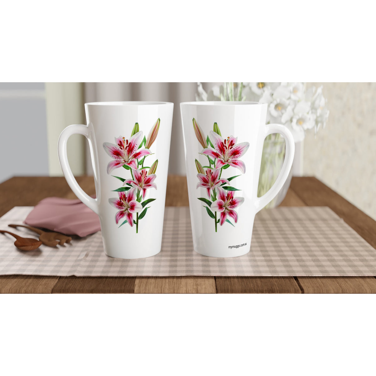 Two tall white ceramic mugs with pink and green lily floral prints, displayed side by side on a checkered placemat over a wooden table.