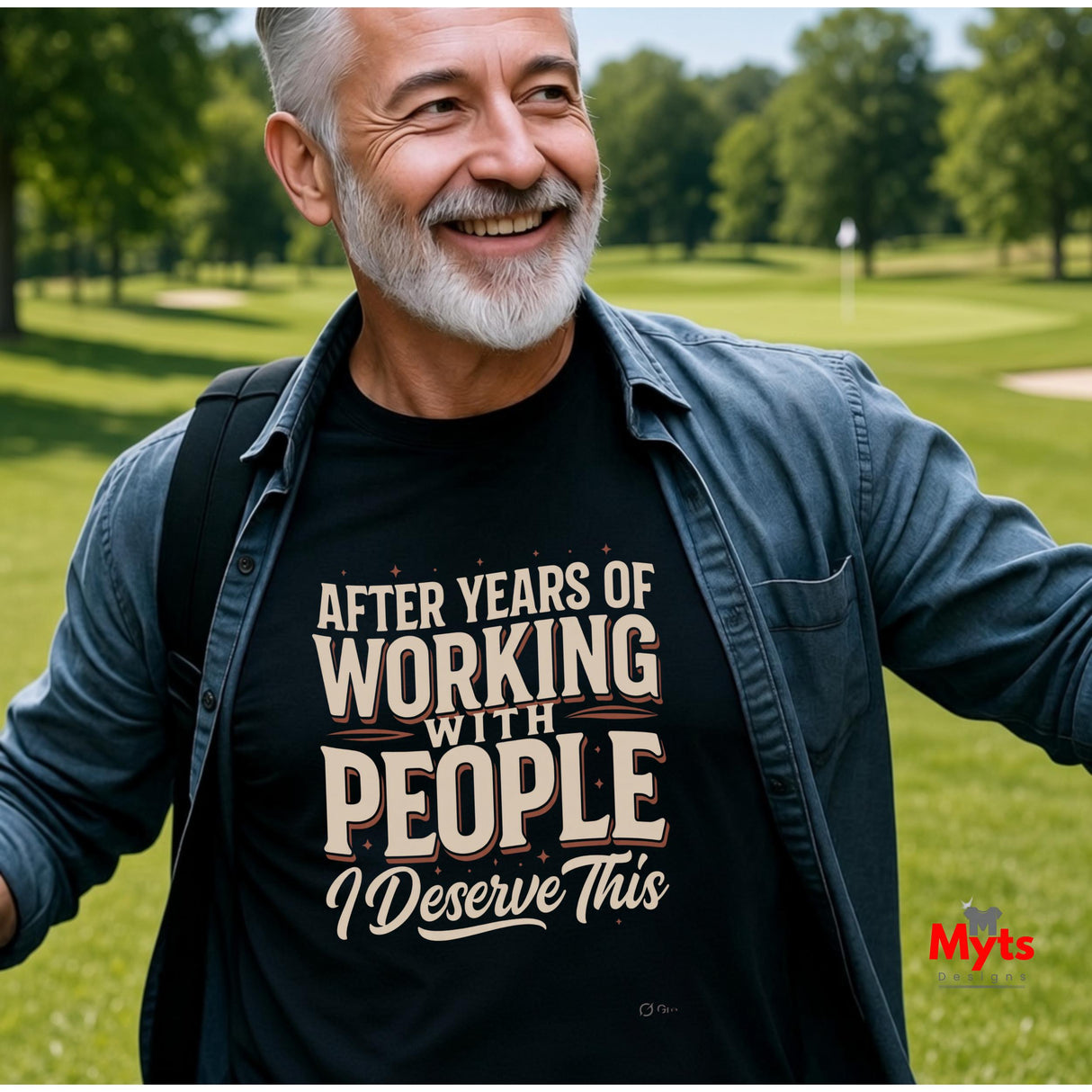 A smiling older man with gray hair and a beard stands outdoors on a grassy golf course wearing a black graphic t-shirt under an open blue denim shirt. The t-shirt features the text 'AFTER YEARS OF WORKING WITH PEOPLE I Deserve This' in beige and white lettering.