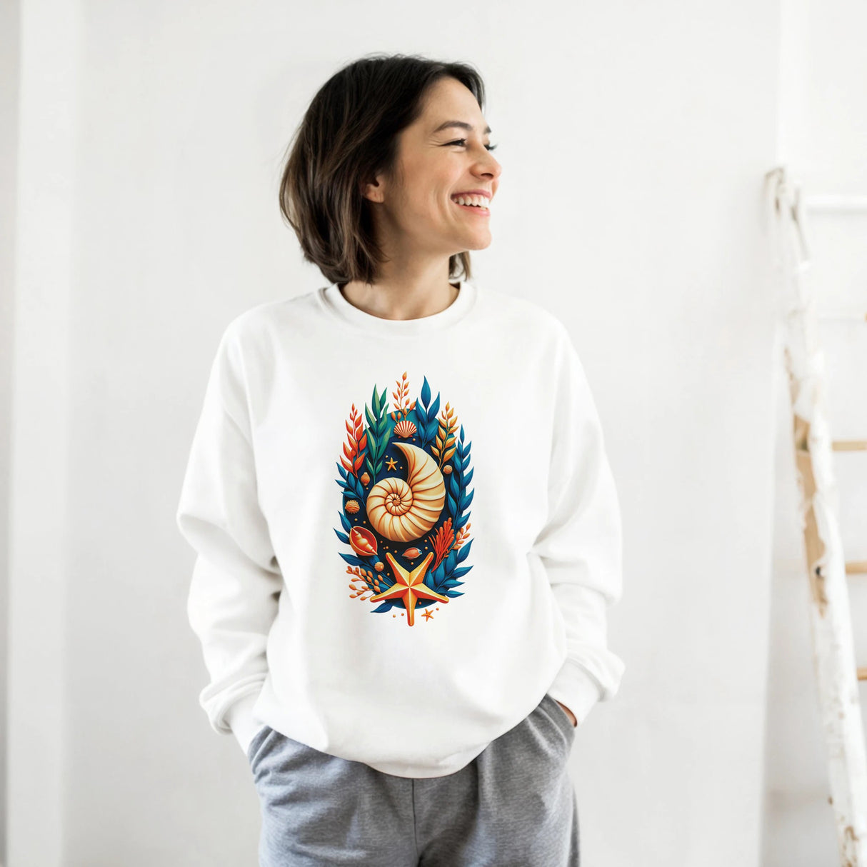 A woman wearing a white crewneck sweatshirt with a colorful seashell and marine life graphic on the front, paired with gray sweatpants, standing against a white wall.