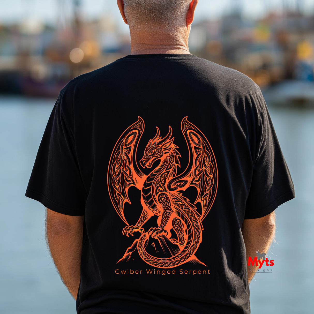 A man viewed from behind wearing a black t-shirt with a large orange graphic of a winged serpent dragon and the text 'Gwiber Winged Serpent' on the back, set against a blurred waterfront background.