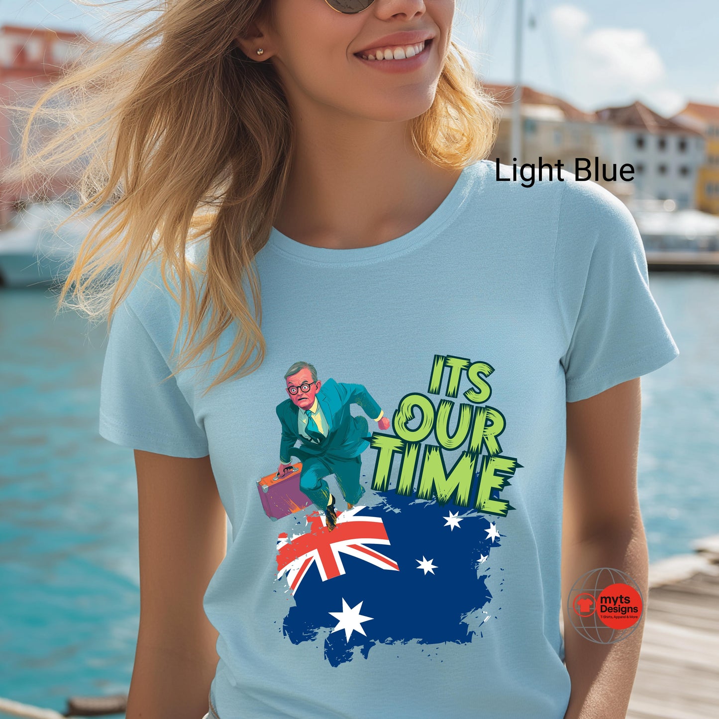 Person wearing a light blue t-shirt with a graphic design and text, standing by a waterfront.