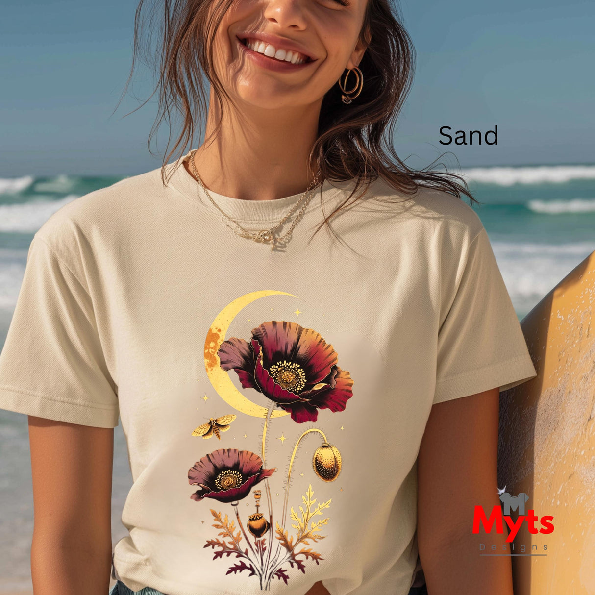 A woman wearing a cream-colored t-shirt with a graphic print of poppy flowers and a crescent moon on a beach. The text 'Sand' indicates the colorway, and a red 'Myts' brand logo is visible in the bottom right corner.