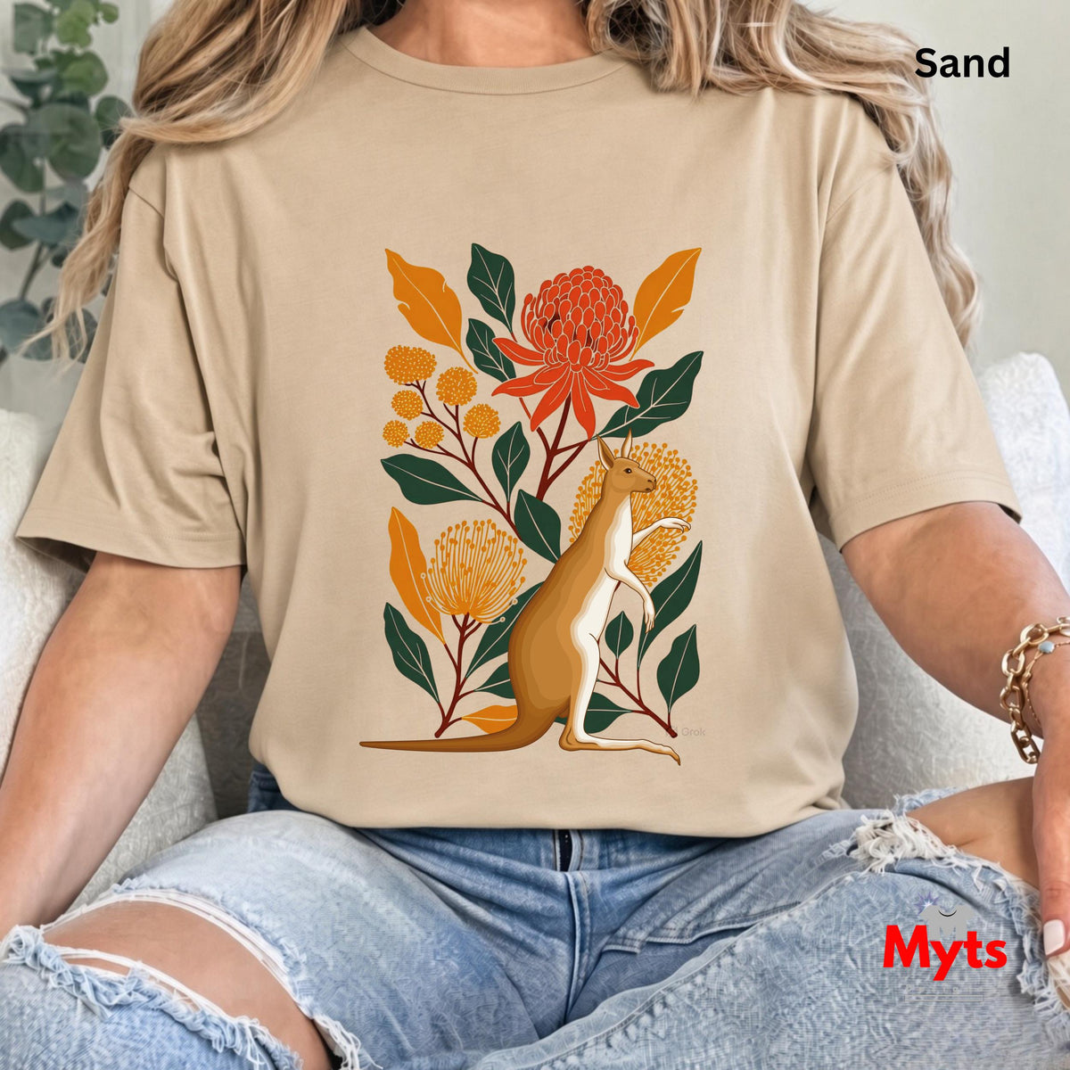 A woman wearing a Sand graphic t-shirt with a kangaroo and floral design, paired with ripped blue jeans, sitting on a white couch. The text 'Sand' is visible in the top right corner.