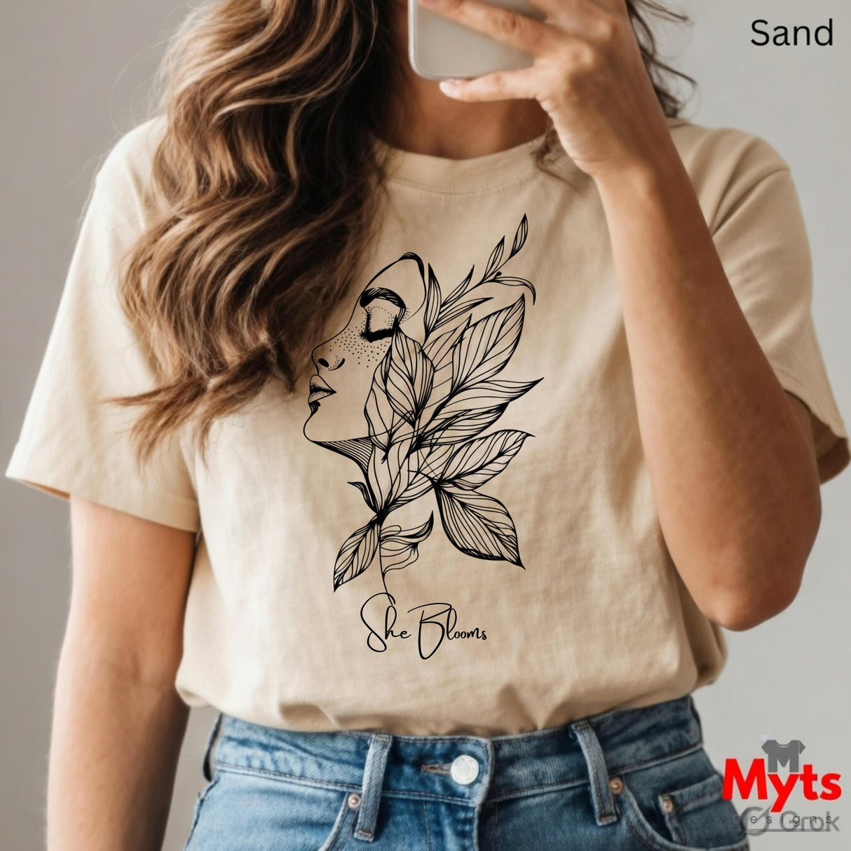 Person wearing a Sand t-shirt with a floral design and text, holding a phone.