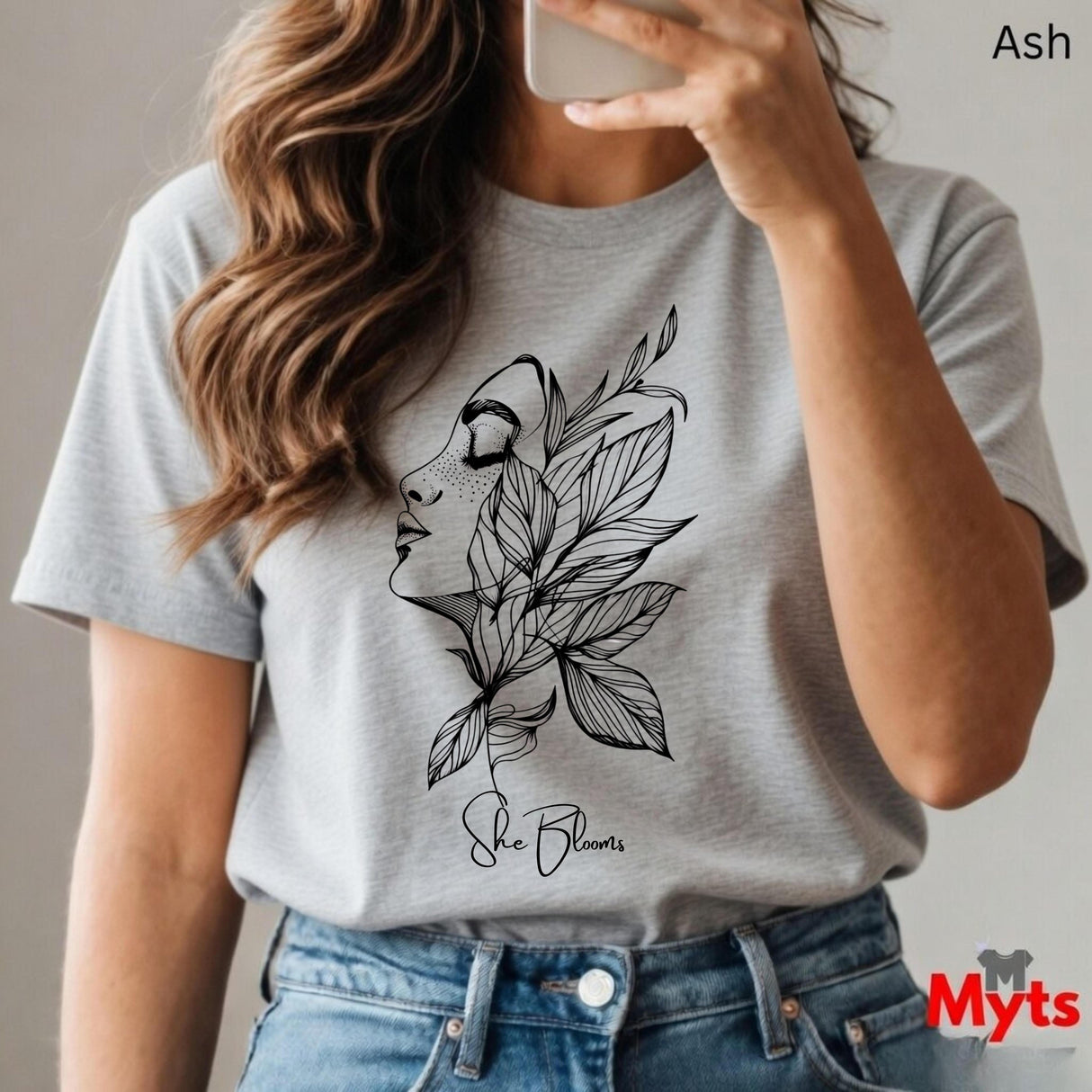 Person wearing a Ash t-shirt with a leaf design and 'She Blooms' text, holding a phone.