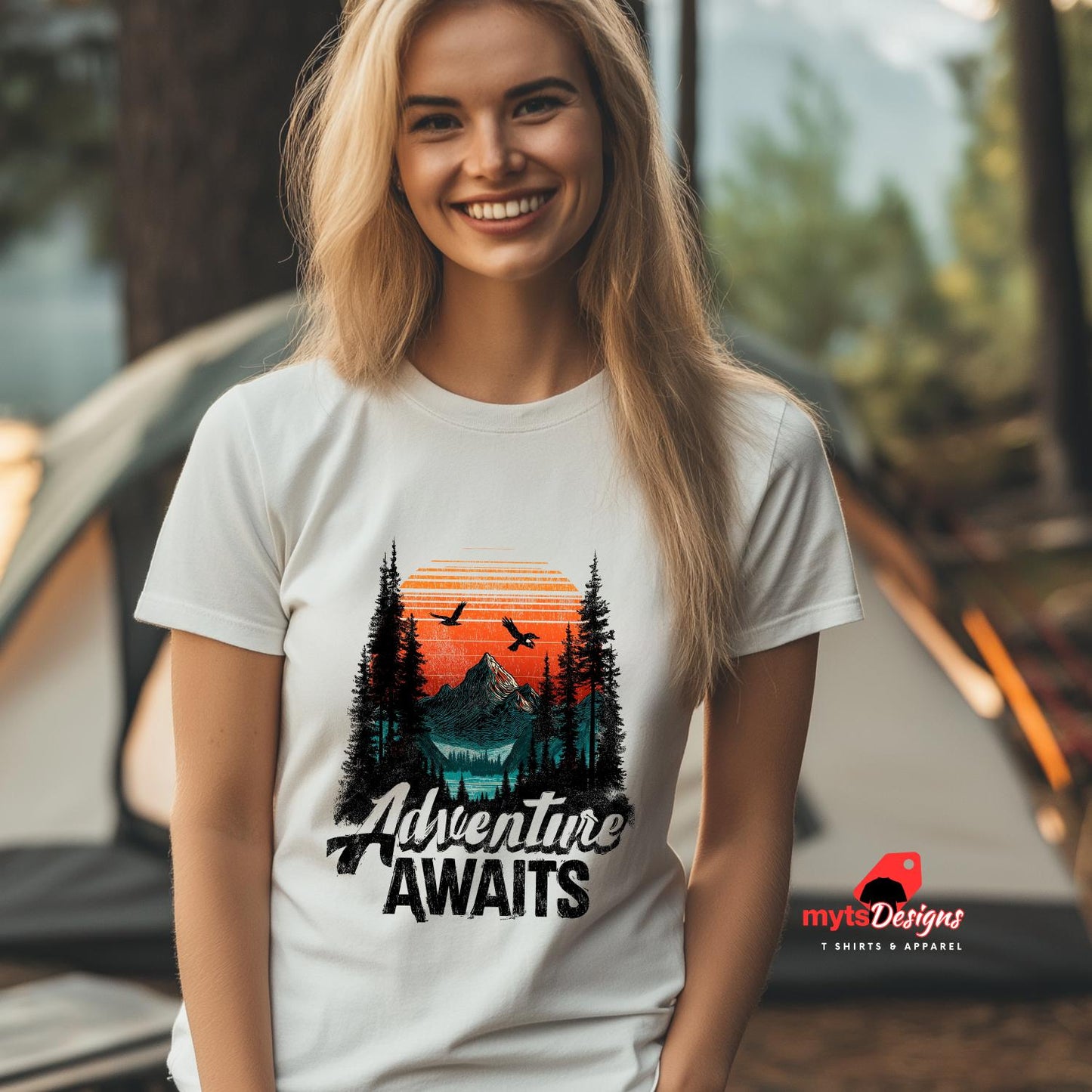 Woman wearing a 'Adventure Awaits' t-shirt with a scenic design, standing in front of a tent.
