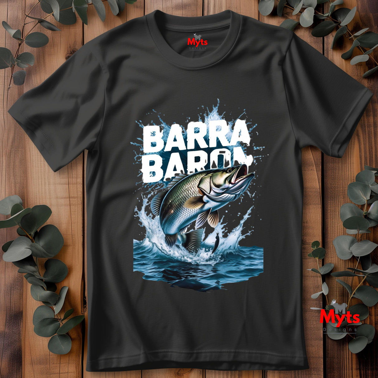Black t-shirt laid flat on wooden surface with “BARRA BARON” text and large realistic jumping barramundi fish graphic splashing out of blue water with fishing lure