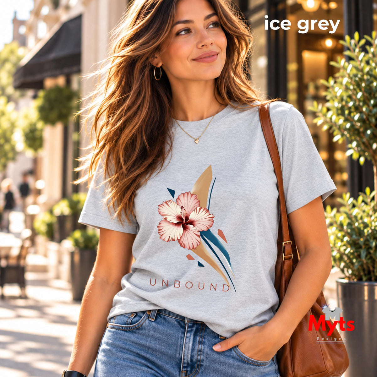 Woman wearing an Ice Grey UNBOUND hibiscus graphic T-shirt with blue jeans on a sunlit shopping street.