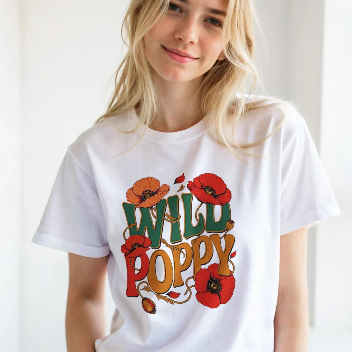A blonde woman wearing a white graphic t-shirt with a colorful retro-style 'WILD POPPY' design, paired with brown pants, standing in a bright, minimalist room.