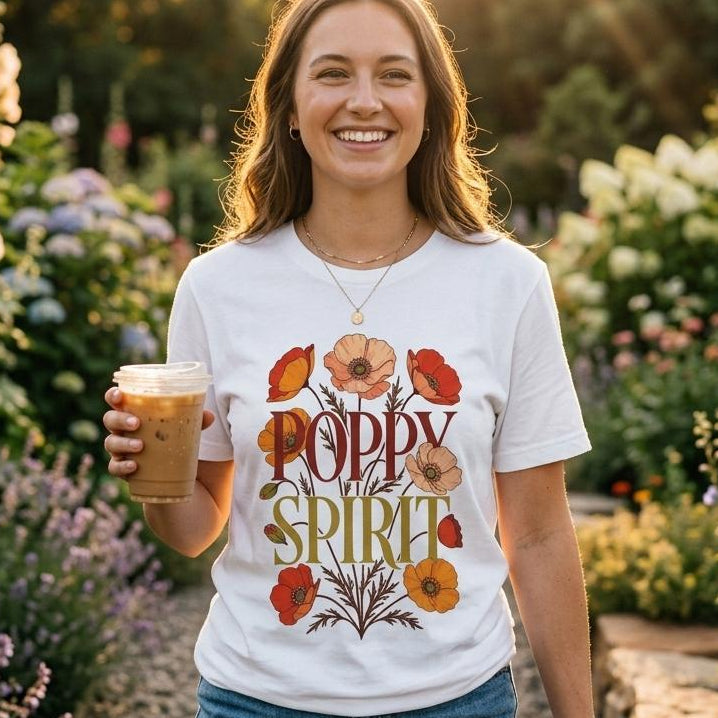 A smiling woman stands in a sunlit flower garden holding an iced coffee. She is wearing a white graphic T-shirt with the words “Poppy Spirit” in warm multicolor lettering surrounded by red, orange, and yellow flowers, paired with blue jeans.