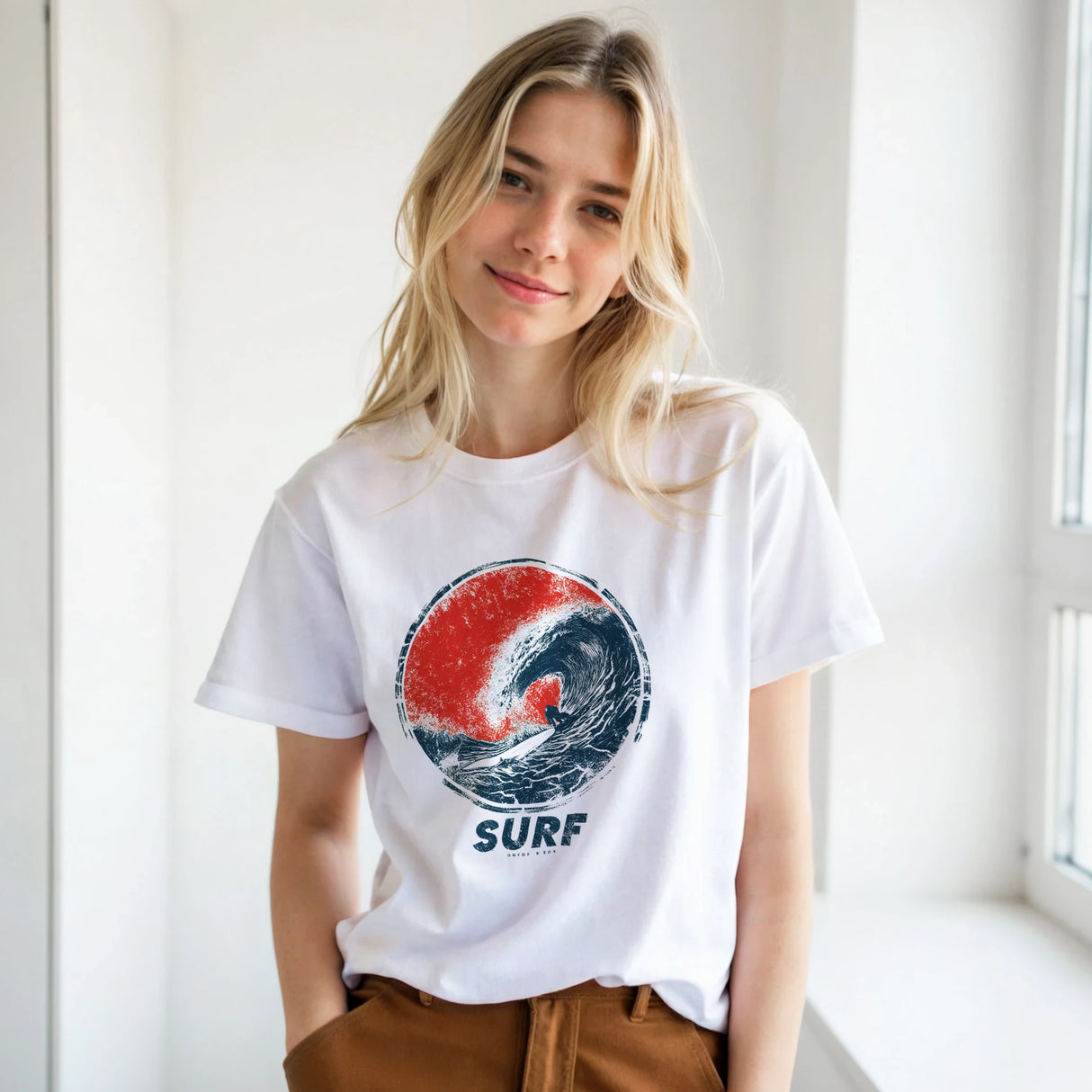 A blonde woman wearing a white graphic t-shirt with a red and blue wave design and the word 'SURF' underneath, paired with brown pants, standing in a bright indoor setting.