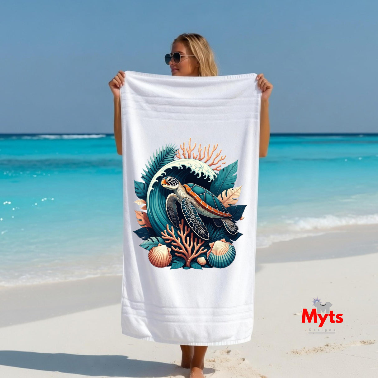 A woman on a sandy beach holds up a large white beach towel featuring a colorful graphic of a sea turtle, coral, and seashells. The background shows a turquoise ocean and blue sky.