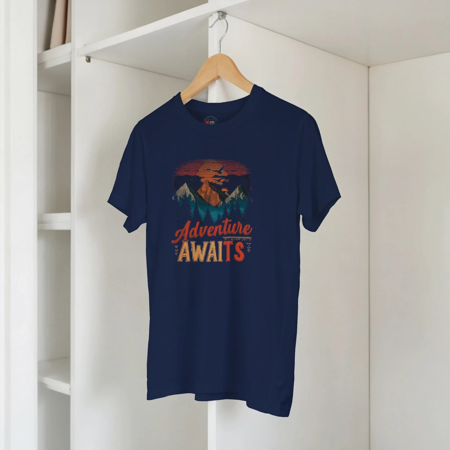 Navy blue t-shirt with 'Adventure Awaits' graphic on a hanger against a white wall.