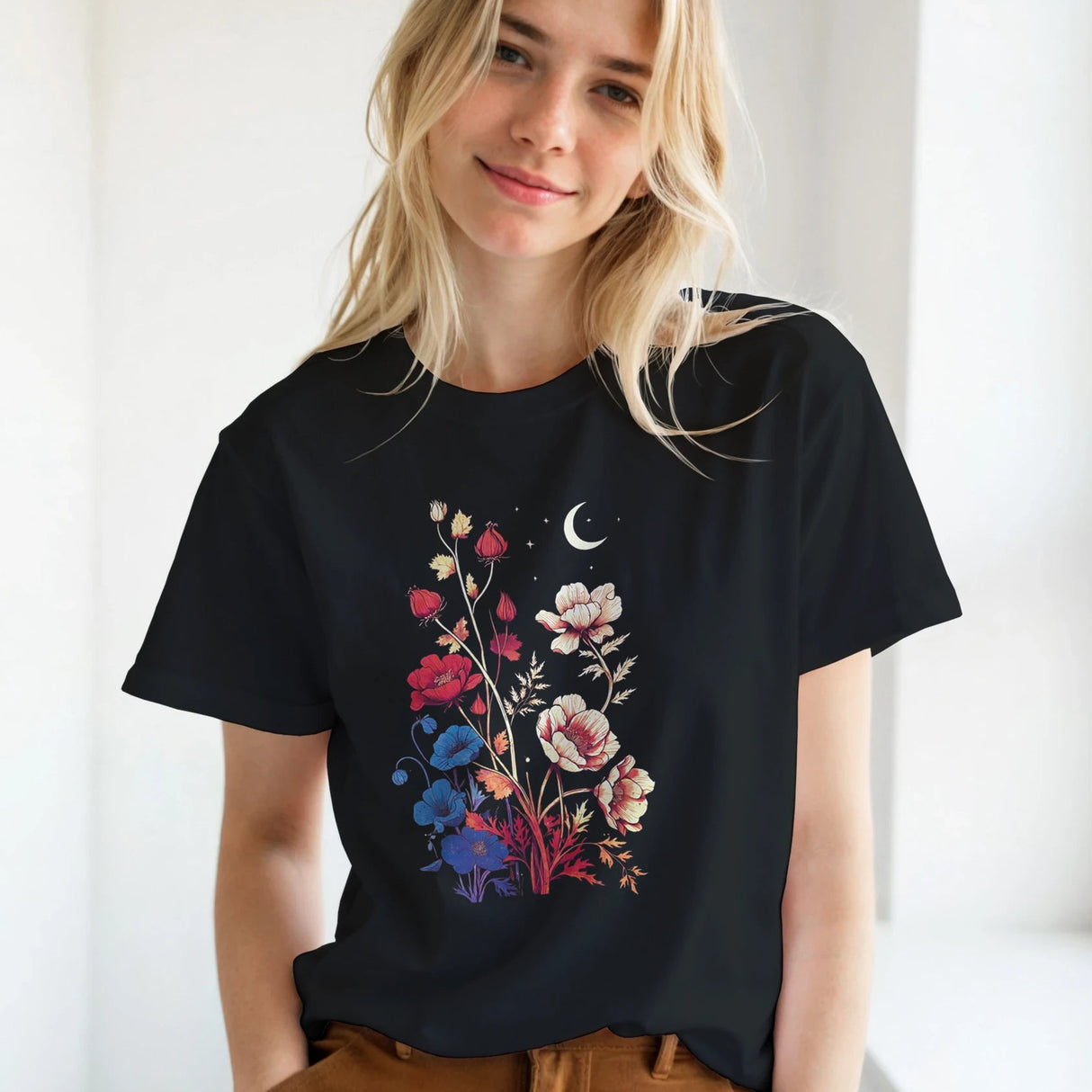 A blonde woman wearing a black short-sleeve t-shirt with a colorful floral and crescent moon graphic, paired with brown pants, standing in a bright indoor setting.