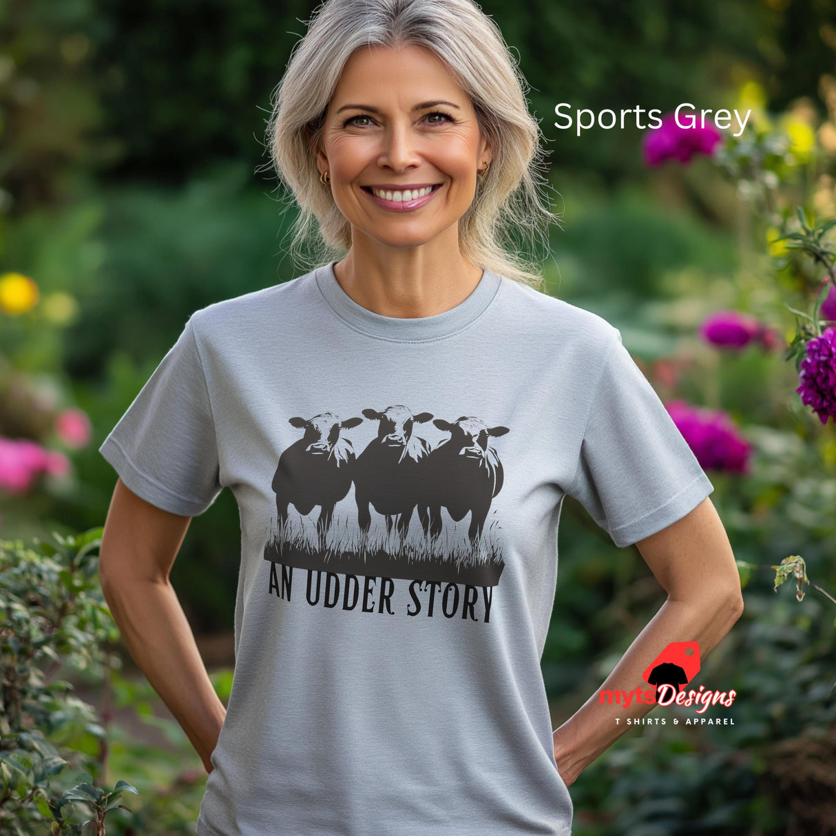 A woman wearing a sports grey cotton T-shirt featuring illustrated cows and text “An Udder Story” – humorous farm-themed graphic tee standing in a cottage garden