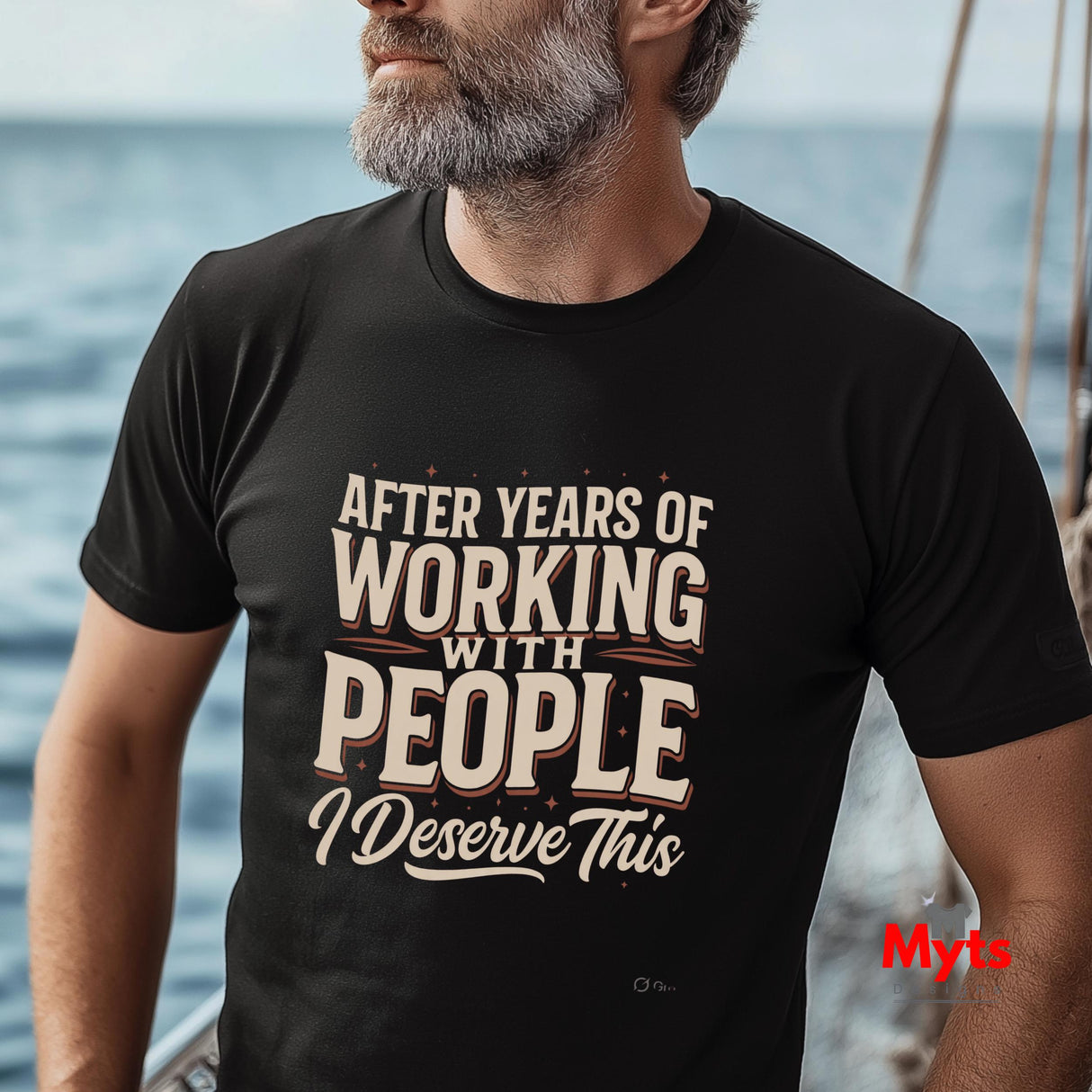 A man wearing a black graphic t-shirt with the text 'AFTER YEARS OF WORKING WITH PEOPLE I DESERVE THIS' in cream and brown lettering, set against a blurred background of water and a boat.