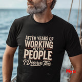 A man wearing a black graphic t-shirt with the text 'AFTER YEARS OF WORKING WITH PEOPLE I DESERVE THIS' in cream and brown lettering, set against a blurred background of water and a boat.