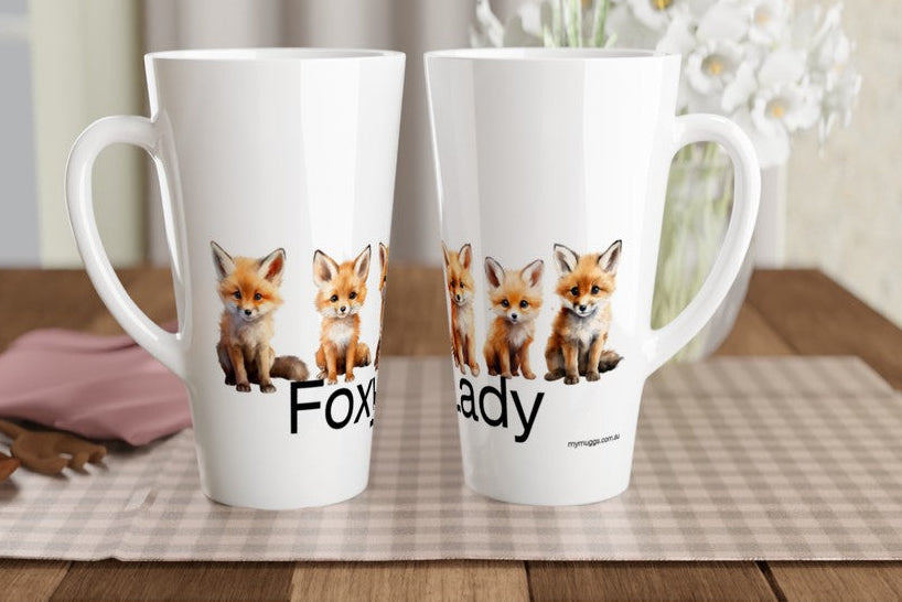 Two white ceramic mugs with printed fox illustrations and partial text, displayed on a checkered placemat on a wooden table with a blurred indoor background.