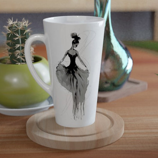 A white ceramic latte mug with a black ink illustration of a ballerina, displayed on a round wooden coaster on a wooden table next to a small potted cactus.