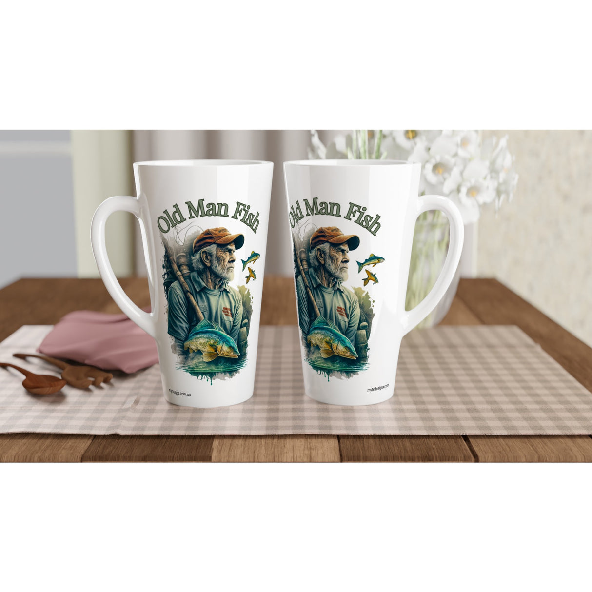Two white ceramic coffee mugs with a printed illustration of an old man fishing and the text 'Old Man Fish', displayed on a checkered table mat on a wooden table.