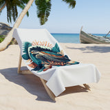 A white beach towel featuring a colorful graphic print of a tropical fish and coral, draped over a wooden lounge chair on a sandy beach with palm trees and the ocean in the background.