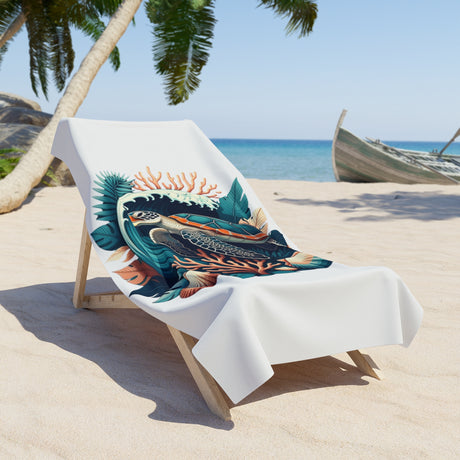 A white beach towel featuring a colorful graphic print of a tropical fish and coral, draped over a wooden lounge chair on a sandy beach with palm trees and the ocean in the background.