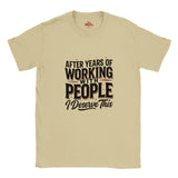 A sand crewneck T-shirt with a humorous graphic printed on the front. The text reads 'AFTER YEARS OF WORKING WITH PEOPLE I Deserve This' in black and dark brown lettering.