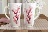 Two tall white ceramic mugs with pink and red cherry blossom branch illustrations, displayed on a checkered placemat on a wooden table with a blurred floral background.