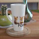 A white ceramic latte mug with a printed graphic of two foxes and the word 'FOX' sits on a round wooden coaster on a wooden table, styled with a small potted cactus and a green plant in a home setting.