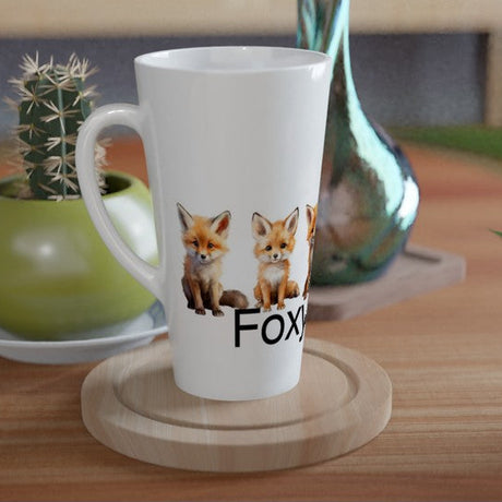 A white ceramic latte mug with a printed graphic of two foxes and the word 'FOX' sits on a round wooden coaster on a wooden table, styled with a small potted cactus and a green plant in a home setting.