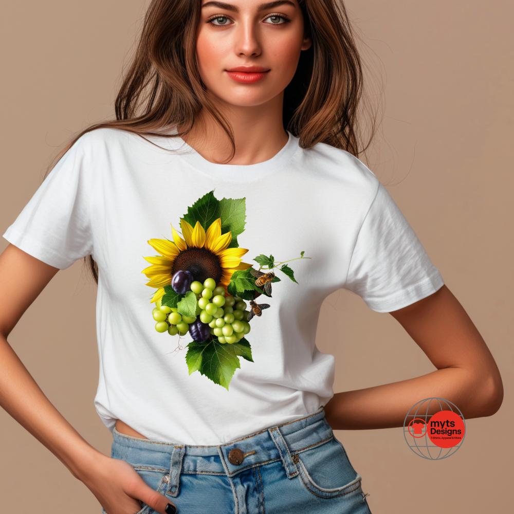 A young woman wears a white t-shirt featuring a vibrant graphic of a sunflower surrounded by green leaves, green and purple grapes, and small twigs. The design is positioned centrally on the shirt, creating a fresh, natural aesthetic. She poses confidently with her hands in her denim jeans pockets against a neutral beige background. The “mytsDesigns” red and black logo appears in the lower corner of the image.