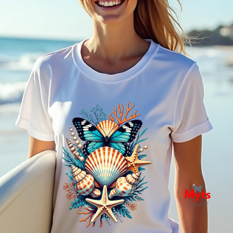 A woman on a beach wearing a white short-sleeve t-shirt with a colorful ocean-themed graphic print featuring a butterfly, seashells, starfish, and coral. A red 'Myts' watermark is visible on the bottom right.