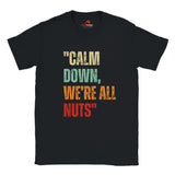 A flat lay view of a black retro styled t-shirt, featuring distressed lettering "Calm Down, We're All Nuts" on a white background
