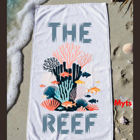 A white beach towel laid flat on sandy shore with ocean foam at the left edge. The towel features the words “THE REEF” in large gray letters surrounding a colorful coral reef and fish illustration.