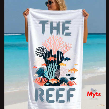 A woman on a beach holds up a large white beach towel featuring a colorful coral reef illustration and the words 'THE REEF' in gray. The background shows a sunny beach with turquoise water and sand.