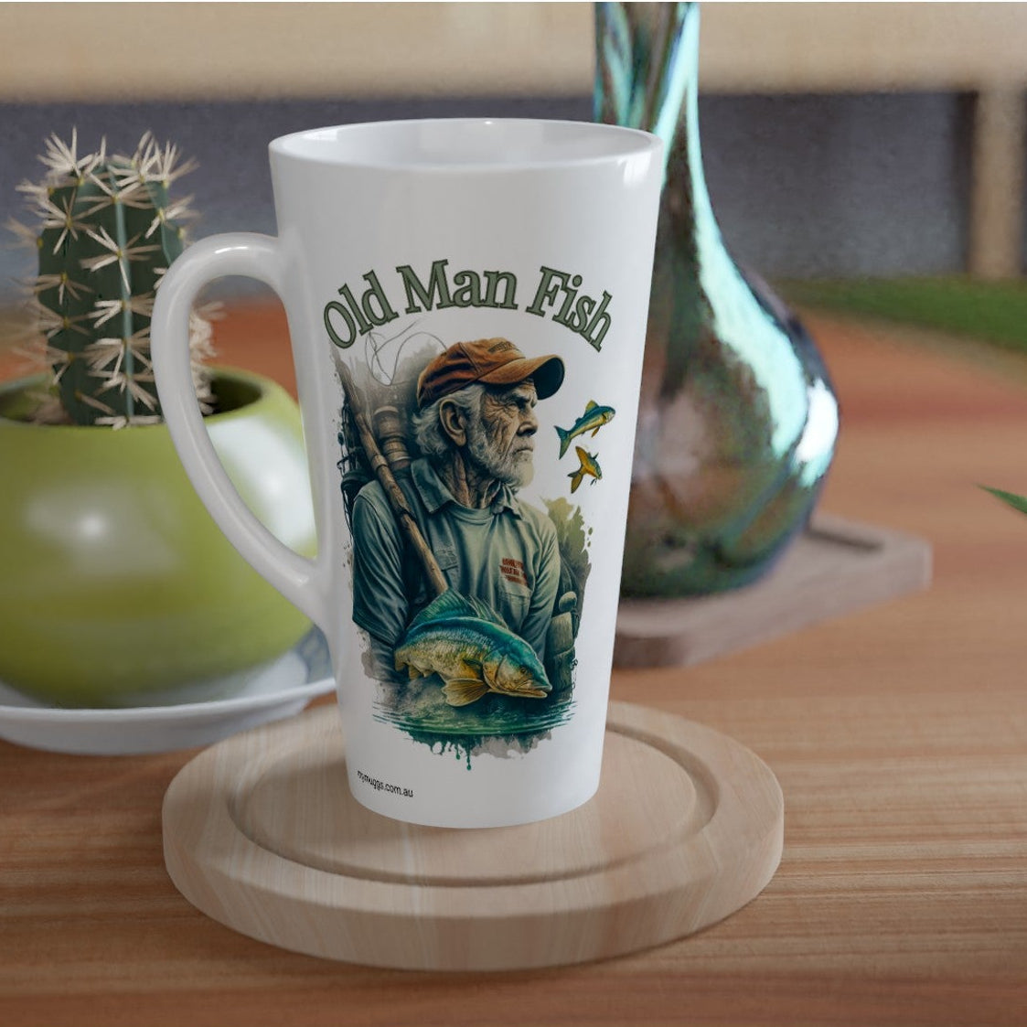 A white ceramic mug with a colorful 'Old Man Fish' illustration sits on a round wooden coaster on a wooden table, styled with a small potted cactus and a metallic vase in the background.