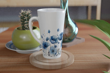 A tall white ceramic mug decorated with a blue poppy floral pattern, resting on a round wooden coaster on a wooden table. A small green cactus in a green pot and a metallic teal vase are visible in the background.
