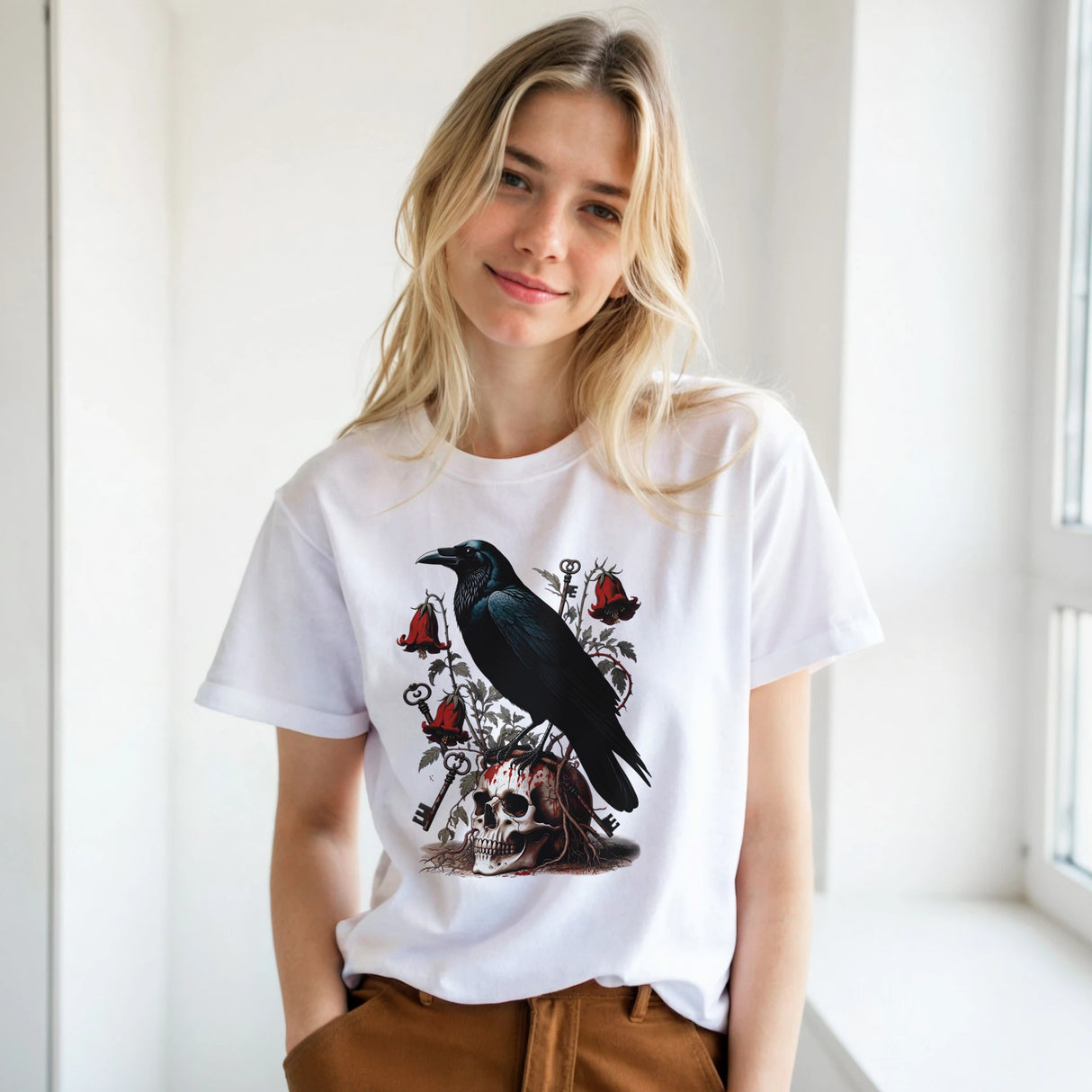 A blonde woman wearing a white graphic t-shirt with a black raven, skull, and red roses design, paired with brown pants, standing in a bright indoor setting.