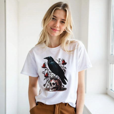 A blonde woman wearing a white graphic t-shirt with a black raven, skull, and red roses design, paired with brown pants, standing in a bright indoor setting.
