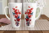 Two tall white ceramic mugs with red and blue floral and butterfly designs, displayed on a checkered placemat on a wooden table.
