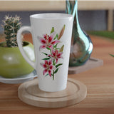 A white ceramic latte mug with a pink and green lily floral print, resting on a round wooden coaster on a wooden table. In the background, a small green cactus in a green pot and a metallic teal glass vase are visible.