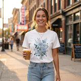A woman smiling on a city sidewalk at sunset, wearing a white graphic T-shirt with a blue floral print and light blue jeans, holding an iced coffee.
