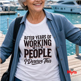 A smiling older woman at a marina wears a white graphic T-shirt with the text 'AFTER YEARS OF WORKING WITH PEOPLE I Deserve This' in black and brown lettering, layered under a light blue denim shirt. Boats and water are visible in the background.