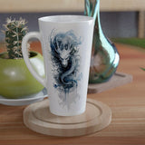 A tall white ceramic mug with a blue dragon illustration, resting on a round wooden coaster on a wooden table. A small green potted cactus and a metallic teal vase are visible in the background.