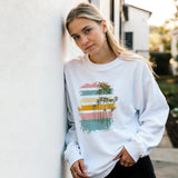 A woman wearing a white sweatshirt with a colorful palm tree design is outdoors.