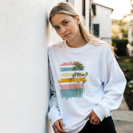 A woman wearing a white sweatshirt with a colorful palm tree design is outdoors.